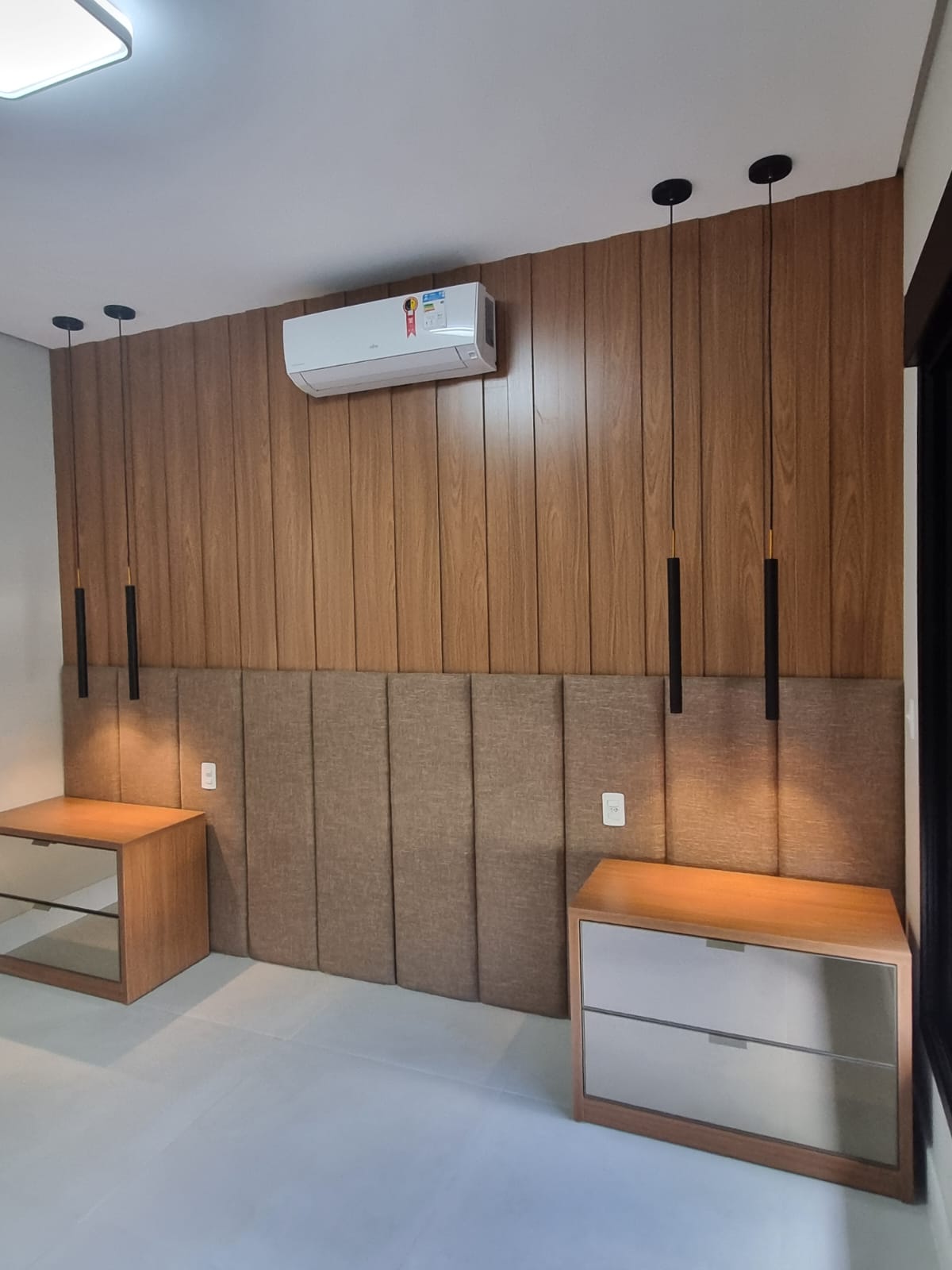 Parede de dormitório com painel ripado amadeirado, cabeceira estofada em tecido e dois criados-mudos com gavetas espelhadas e pendentes pretos